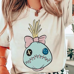 May include: Cream-colored t-shirt featuring a cartoon voodoo doll design. The doll has a light blue body, a pink bow, and yellow hair. It has a black cross for an eye and a blue button eye. The doll is stitched around the edges.