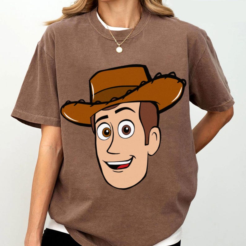 May include: Brown t-shirt featuring a cartoon illustration of a smiling cowboy character wearing a brown hat. The graphic is centered on the front of the shirt. The shirt has short sleeves and a crew neck.