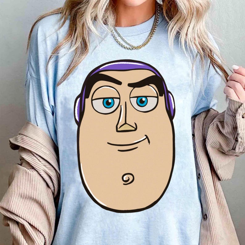 May include: Light blue t-shirt featuring a cartoon illustration of Buzz Lightyear's face. The design includes his signature purple hair, blue eyes, and a confident smirk. The shirt is a casual wear item, perfect for fans of the animated movie.