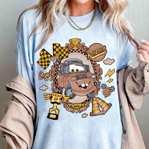 May include: Light blue t-shirt featuring a cartoon image of Mater from the Cars movie. The design includes a brown Mater character inside a decorative frame, along with racing flags, a trophy, and the words "Rusteze" and "Piston Cup".