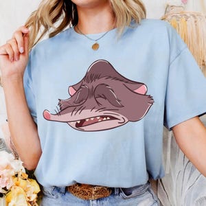 Peut inclure: T-shirt bleu clair avec une illustration de dessin anim&eacute; d'un rongeur grincheux. Le rongeur a les yeux ferm&eacute;s et la bouche ouverte, dans des tons de marron et de rose. Le t-shirt a des manches courtes et un col rond.