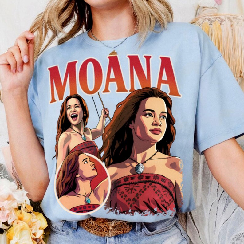 以下が含まれることがあります： ライトブルーのTシャツで、キャラクター「モアナ」の赤とオレンジの大きなグラフィックプリントが施されています。柄の入った赤いトップスと青い石のネックレスを身につけたキャラクターの3つの異なるイラストと、上部に「MOANA」の文字がデザインされています。