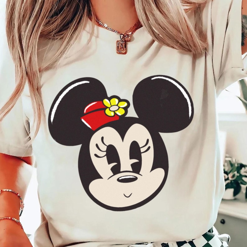 Peut inclure: T-shirt cr&egrave;me avec un grand dessin du visage de Minnie Mouse. Le motif comprend des oreilles noires, un n&oelig;ud rouge avec une fleur jaune et un visage souriant. Le t-shirt est un v&ecirc;tement d&eacute;contract&eacute;.