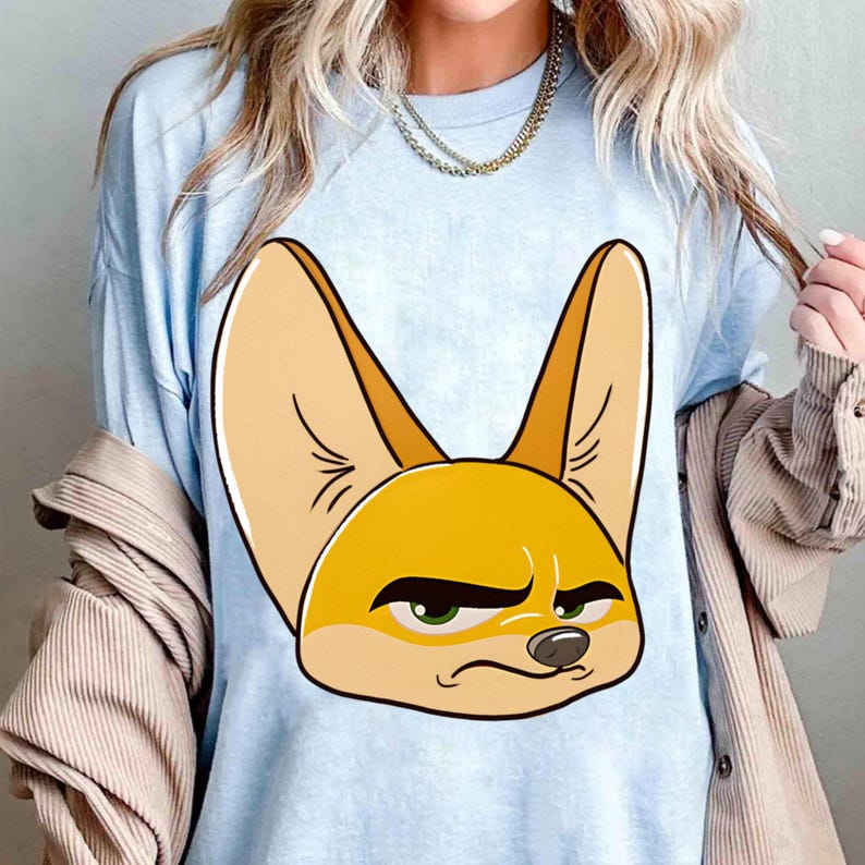 May include: Light blue t-shirt featuring a cartoon illustration of a fennec fox with large ears and a grumpy expression. The fox is primarily yellow and tan with green eyes. The shirt is a solid color and has a crew neck.