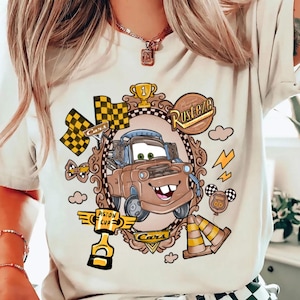 May include: Beige t-shirt featuring a cartoon image of Tow Mater from the movie Cars, framed by a decorative border. The design includes racing flags, a trophy, and the words "Rusteze" and "Cars". Additional graphics include lightning bolts and traffic cones.