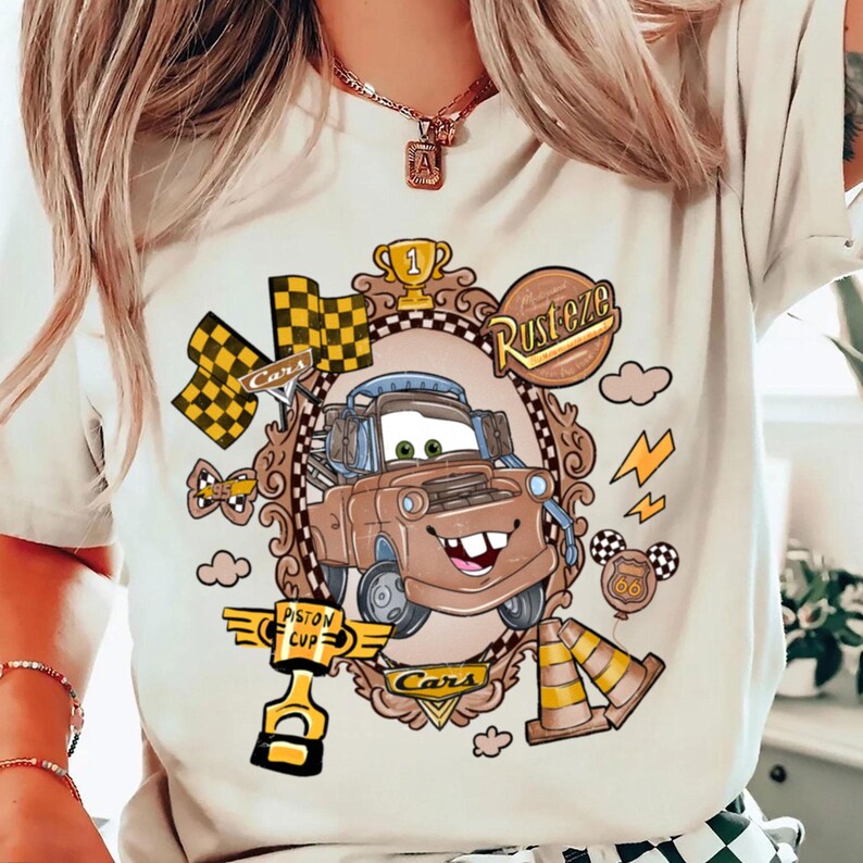 May include: Beige t-shirt featuring a cartoon image of Tow Mater from the movie Cars, framed by a decorative border. The design includes racing flags, a trophy, and the words "Rusteze" and "Cars". Additional graphics include lightning bolts and traffic cones.