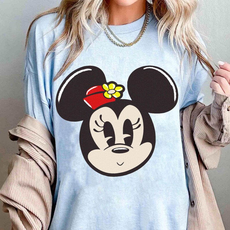Peut inclure: T-shirt bleu clair avec un motif de Minnie Mouse. Le motif repr&eacute;sente le visage de Minnie avec des oreilles noires, un n&oelig;ud rouge avec une fleur jaune et une expression souriante. Le t-shirt est de couleur unie et semble &ecirc;tre fait d'un tissu doux.