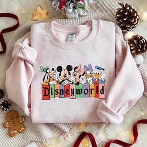 May include: A light pink sweatshirt featuring embroidered Disney characters: Goofy, Mickey Mouse, Minnie Mouse, Daisy Duck, and Donald Duck. The word "Disneyworld" is embroidered in colorful letters below the characters. The sweatshirt is made of a soft material.