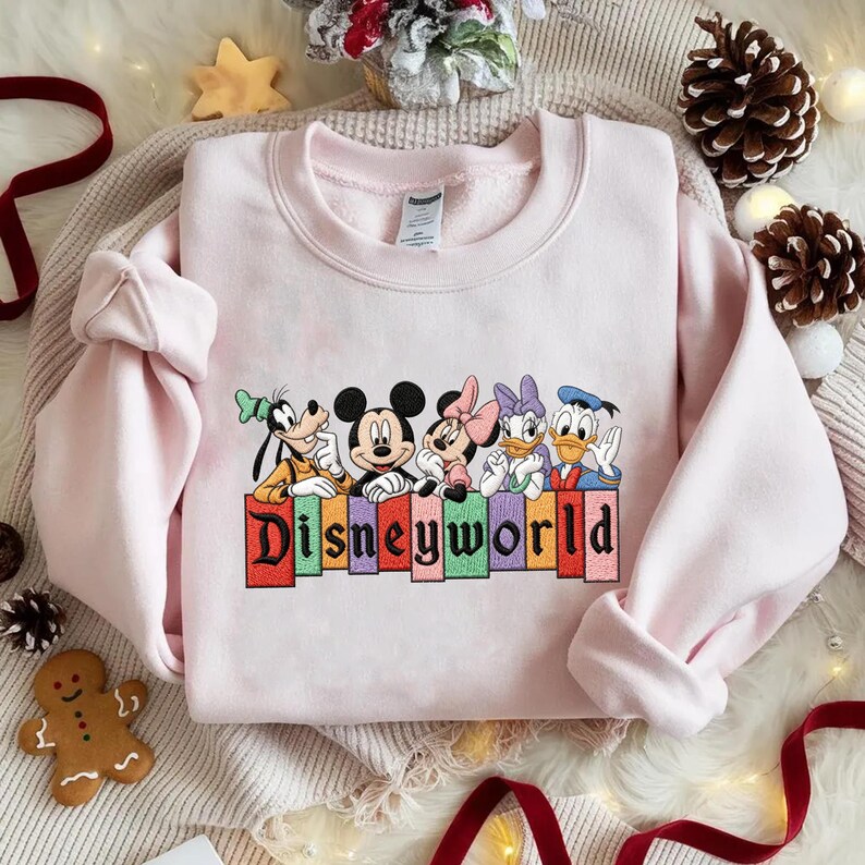 May include: A light pink sweatshirt featuring embroidered Disney characters: Goofy, Mickey Mouse, Minnie Mouse, Daisy Duck, and Donald Duck. The word "Disneyworld" is embroidered in colorful letters below the characters. The sweatshirt is made of a soft material.