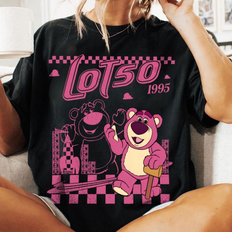 May include: Black t-shirt featuring a pink graphic of Lotso, a bear character, with the text "Lotso 1995". The design includes a checkered pattern and a silhouette of a building. The bear is holding a mallet.