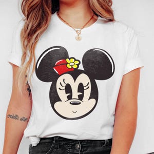 Peut inclure: T-shirt blanc avec un dessin du visage de Minnie Mouse. Le motif comprend des oreilles noires, un n&oelig;ud rouge avec une fleur jaune et un visage souriant. Le t-shirt a des manches courtes.