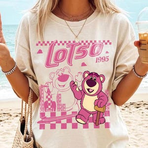 May include: Beige t-shirt featuring a pink graphic of Lotso the bear from Toy Story, with the text "LOTSO 1995". The design includes a checkered border and a cartoon illustration of Lotso holding a mallet.