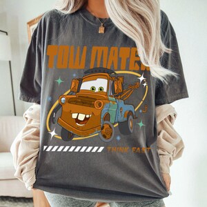 May include: A charcoal gray short-sleeve t-shirt featuring a graphic of the character Tow Mater. The design includes the text "TOW MATER" in orange at the top and "THINK FAST" in orange at the bottom, accented by small star shapes.