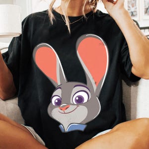 Puede incluir: Camiseta negra con una ilustraci&oacute;n de dibujos animados de un conejo. El conejo es gris con orejas rosas, ojos morados y un cuello azul. El dise&ntilde;o tiene una expresi&oacute;n sonriente. Es una prenda de vestir informal.