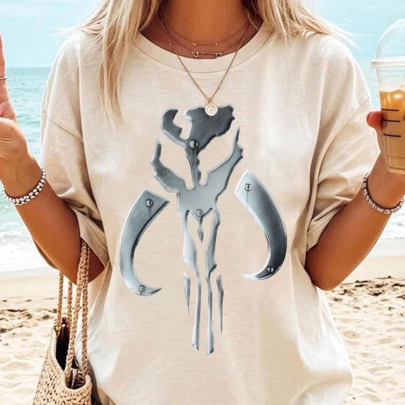 May include: A cream-coloured t-shirt with a metallic silver emblem. The emblem has a symmetrical design with curved elements and rivets. The t-shirt is a casual style, suitable for everyday wear.