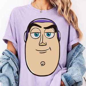 May include: A light purple t-shirt featuring a large cartoon graphic of Buzz Lightyear's face. The graphic has a tan face, blue eyes, and a purple headpiece. The shirt has short sleeves and a relaxed fit.