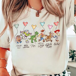 May include: A cream-coloured t-shirt with a colourful illustration of Toy Story characters holding balloon-shaped balloons. Characters include Potato Head, Buzz Lightyear, Rex, Woody, Bullseye, and Bo Peep. Text below each character identifies them.