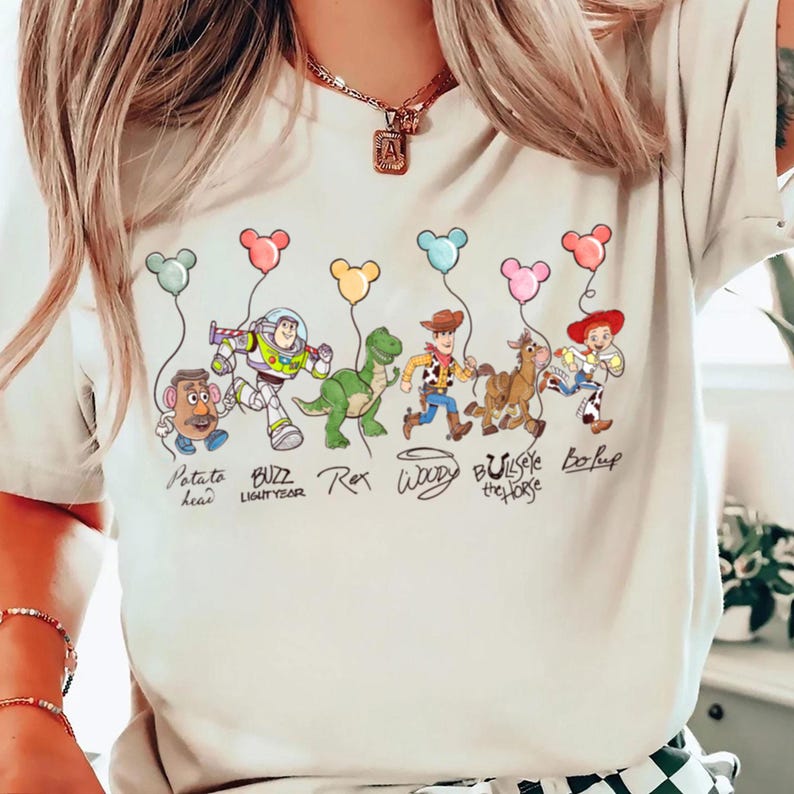 May include: A cream-coloured t-shirt with a colourful illustration of Toy Story characters holding balloon-shaped balloons. Characters include Potato Head, Buzz Lightyear, Rex, Woody, Bullseye, and Bo Peep. Text below each character identifies them.