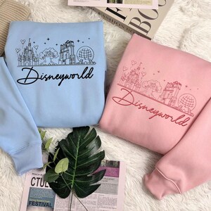 May include: Two sweatshirts, one light blue and one pink, each featuring a black or red "Disneyworld" script and a line drawing of a castle, tree, and globe. The sweatshirts are made of a soft material.