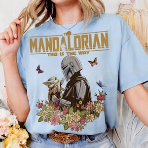 May include: Light blue t-shirt featuring a graphic of The Mandalorian and Grogu surrounded by flowers and butterflies. The text "THE MANDALORIAN" and "THIS IS THE WAY" are printed in gold across the top of the design. A casual, short-sleeved tee.