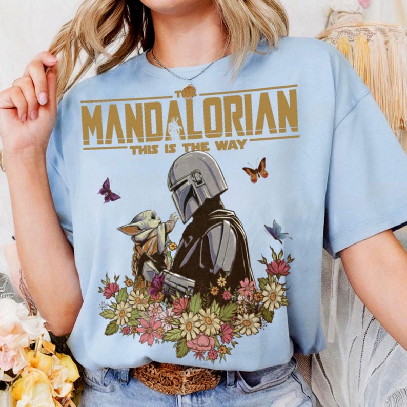May include: Light blue t-shirt featuring a graphic of The Mandalorian and Grogu surrounded by flowers and butterflies. The text "THE MANDALORIAN" and "THIS IS THE WAY" are printed in gold across the top of the design. A casual, short-sleeved tee.