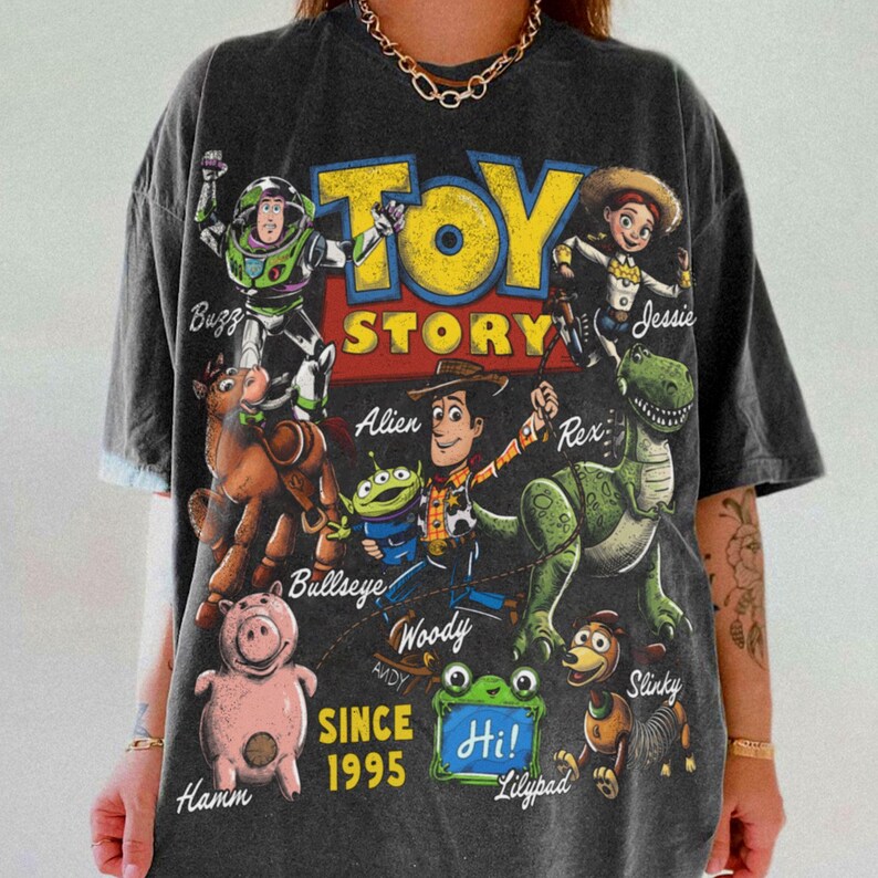 Pode incluir: Uma t-shirt de manga curta cinzento-carv&atilde;o com uma grande impress&atilde;o gr&aacute;fica das personagens de Toy Story, incluindo Buzz Lightyear, Woody, Jessie, Rex, Hamm, Bullseye e um alien&iacute;gena. O texto diz Toy Story, Since 1995 e Hi! Lilypad.