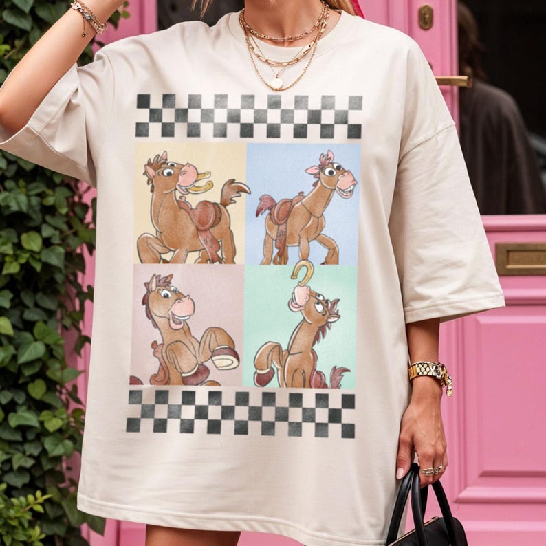 May include: Oversized beige t-shirt featuring a cartoon horse design in four panels with a black and white checkered border. The horse is brown with a saddle and a horseshoe. The background panels are yellow, blue, pink, and green.