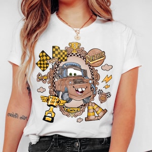 May include: White t-shirt featuring a cartoon image of Mater from the movie Cars. The design includes a brown Mater character, checkered flags, and the words "Cars" and "Rusteze". The shirt has a crew neck and short sleeves.