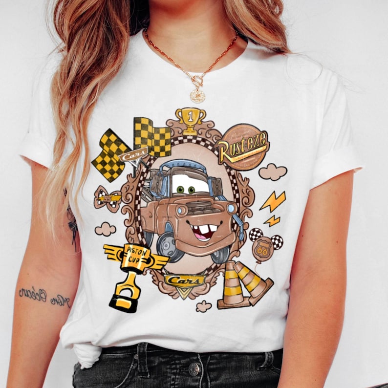 May include: White t-shirt featuring a cartoon image of Mater from the movie Cars. The design includes a brown Mater character, checkered flags, and the words "Cars" and "Rusteze". The shirt has a crew neck and short sleeves.