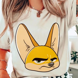 May include: White t-shirt featuring a cartoon illustration of a fennec fox with large ears and a grumpy expression. The fox is primarily yellow and tan with green eyes and black details. The shirt is a casual style.