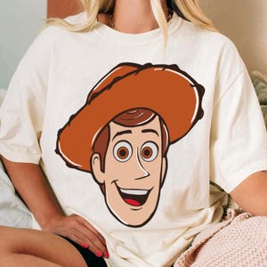 May include: Cream-colored t-shirt featuring a cartoon illustration of a smiling cowboy character wearing a brown hat. The character has brown hair, brown eyes, and a red mouth. The design is centered on the front of the shirt.