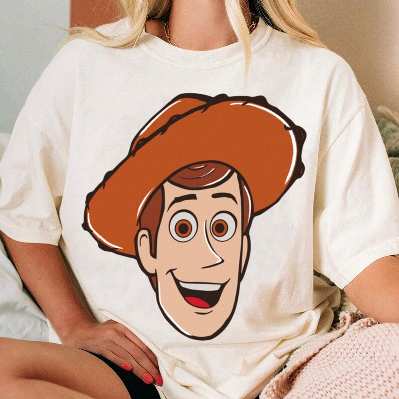 May include: Cream-colored t-shirt featuring a cartoon illustration of a smiling cowboy character wearing a brown hat. The character has brown hair, brown eyes, and a red mouth. The design is centered on the front of the shirt.
