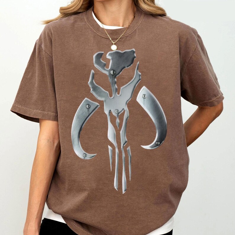 May include: A brown t-shirt with a large, metallic silver emblem. The emblem has a stylized, angular design with curved and pointed elements. The shirt has short sleeves and a crew neck, with a white undershirt visible.