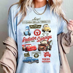 Pu&ograve; includere: Una t-shirt azzurro chiaro con una stampa grafica di personaggi e loghi del film Cars. Il design include testi come Luigi's Casa Della Tires, Flo's Cafe, Radiator Springs Since 1909, Cozy Cone Motel, Fillmore's e Tow Mater Towing.
