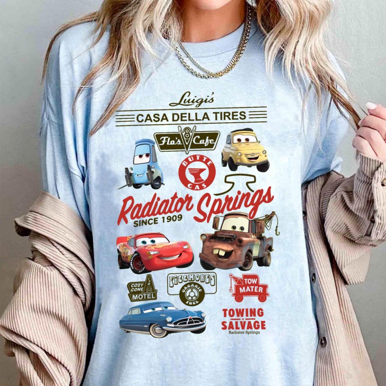 Pu&ograve; includere: Una t-shirt azzurro chiaro con una stampa grafica di personaggi e loghi del film Cars. Il design include testi come Luigi's Casa Della Tires, Flo's Cafe, Radiator Springs Since 1909, Cozy Cone Motel, Fillmore's e Tow Mater Towing.