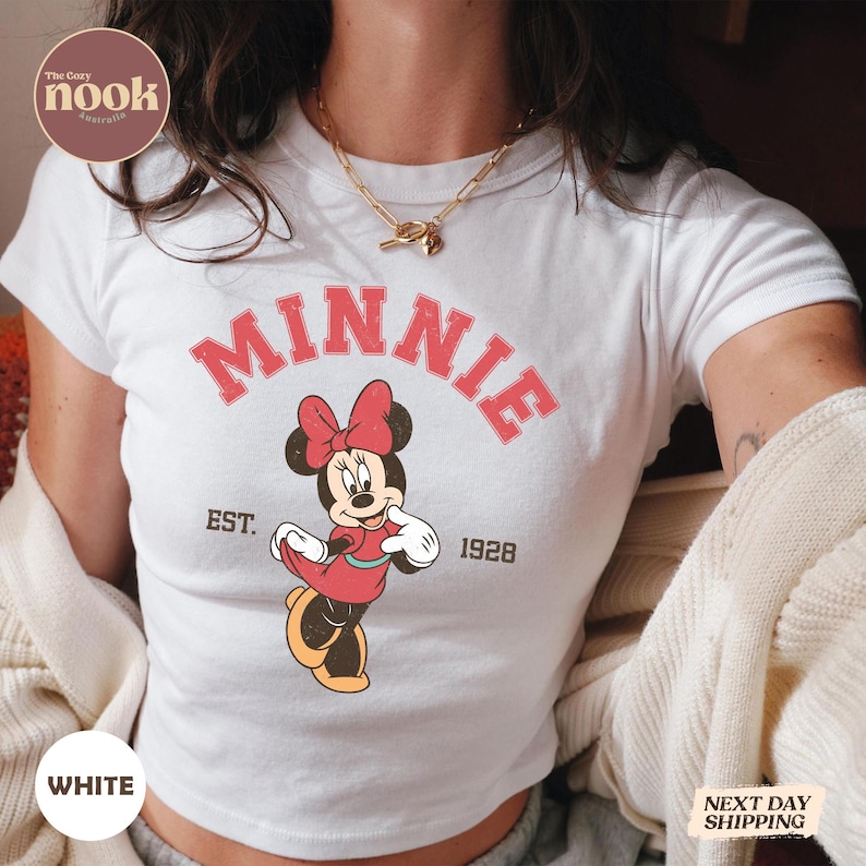 May include: White t-shirt featuring a graphic of Minnie Mouse in a red dress and bow, with the word "MINNIE" in red arched text above her. The shirt also includes "EST. 1928".