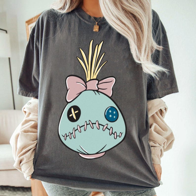 May include: Dark gray t-shirt featuring a cartoon doll design. The doll has a light blue face with a pink bow, yellow hair, and stitched details. The shirt is a comfortable, oversized fit.