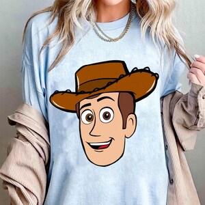 May include: Light blue t-shirt featuring a cartoon illustration of Woody from Toy Story. The graphic shows Woody's face, including his brown cowboy hat, brown hair, and smiling expression. The shirt is a solid light blue color.