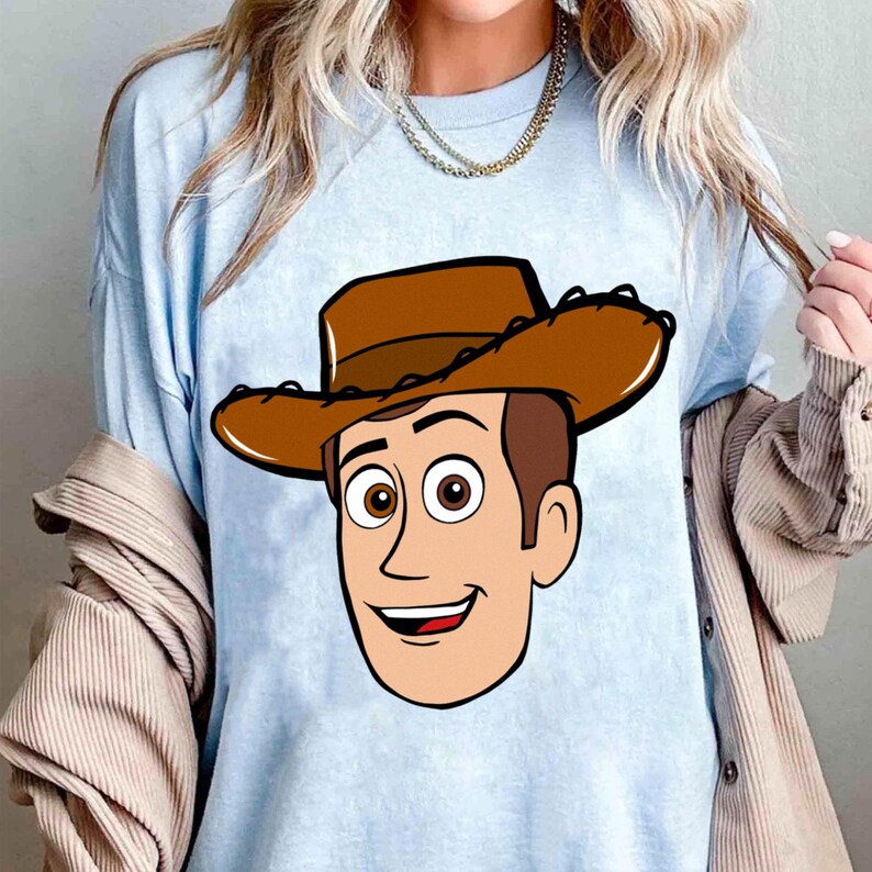 May include: Light blue t-shirt featuring a cartoon illustration of Woody from Toy Story. The graphic shows Woody's face, including his brown cowboy hat, brown hair, and smiling expression. The shirt is a solid light blue color.