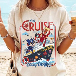 May include: Beige t-shirt featuring a colorful graphic of a cruise ship with cartoon characters. The design includes the text "Let's Cruise" and "Disney Destiny". The ship has a red, white, and blue color scheme.