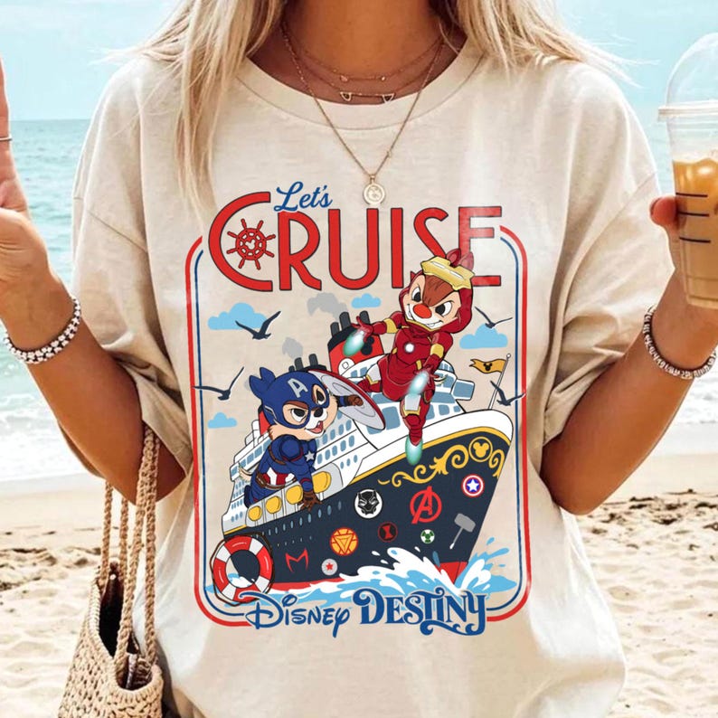 May include: Beige t-shirt featuring a colorful graphic of a cruise ship with cartoon characters. The design includes the text "Let's Cruise" and "Disney Destiny". The ship has a red, white, and blue color scheme.