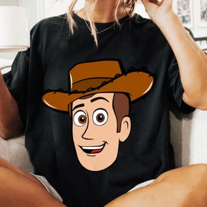 May include: Black t-shirt featuring a cartoon illustration of a cowboy character with a brown hat and smiling face. The design is centered on the shirt, showcasing the character's features.