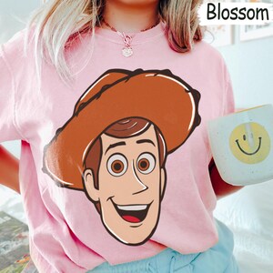 May include: Pink sweatshirt featuring a large cartoon graphic of a cowboy character with a brown hat and smiling face. The sweatshirt also has the word "Blossom" printed on the top right corner. A white mug with a yellow smiley face is held in the hand.