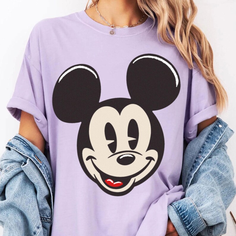 May include: A light purple t-shirt featuring a large graphic of Mickey Mouse's face. The cartoon character's face is black and white with a red mouth. The shirt has short sleeves and a relaxed fit.