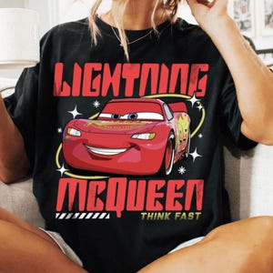 May include: A black t-shirt featuring a graphic of the red race car Lightning McQueen. The text reads LIGHTNING MCQUEEN in large red letters above and below the car, with the words THINK FAST in smaller yellow text at the bottom of the design.