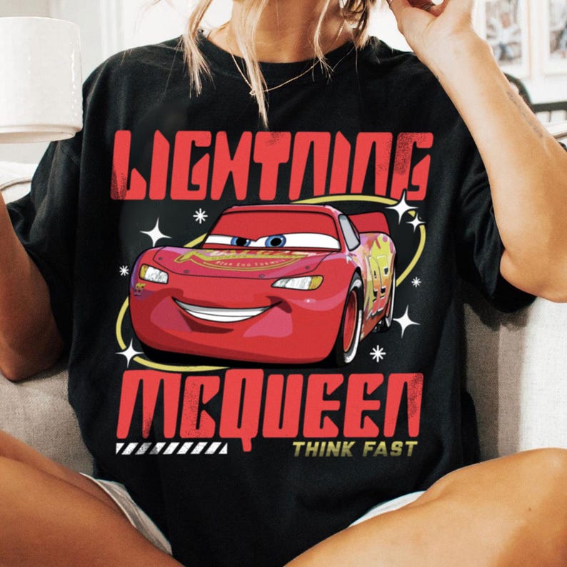 May include: A black t-shirt featuring a graphic of the red race car Lightning McQueen. The text reads LIGHTNING MCQUEEN in large red letters above and below the car, with the words THINK FAST in smaller yellow text at the bottom of the design.