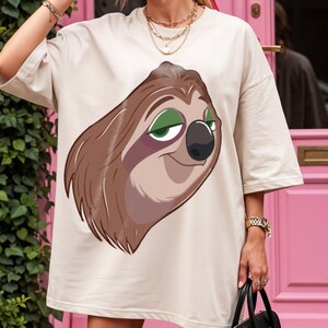May include: Cream-colored t-shirt featuring a cartoon sloth with brown fur, green eyes, and a black nose. The sloth is smiling. The shirt has short sleeves.