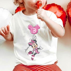 May include: A white ceramic mug with a black handle and the text "But First, Coffee" in a bold, black font. The mug is set against a blurred background. The mug appears to be approximately 10 cm tall.