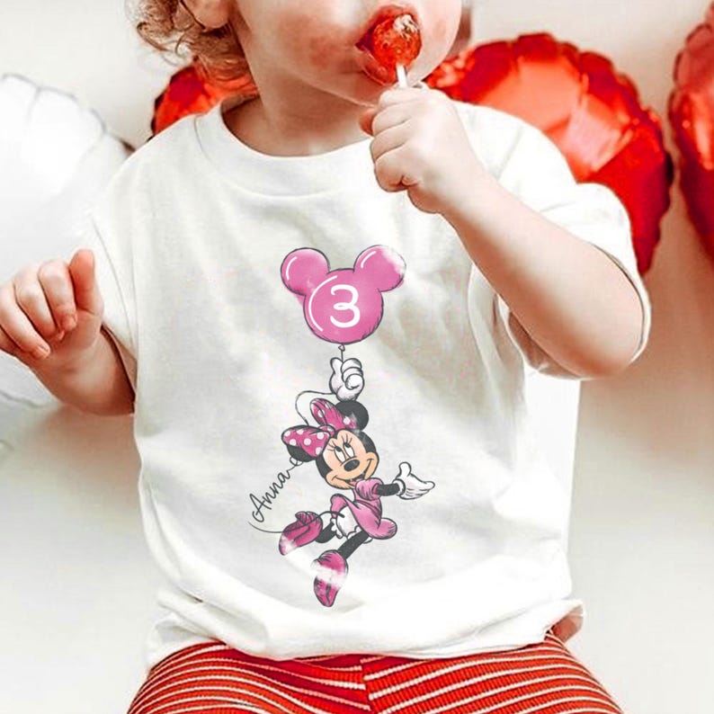 May include: A white ceramic mug with a black handle and the text "But First, Coffee" in a bold, black font. The mug is set against a blurred background. The mug appears to be approximately 10 cm tall.