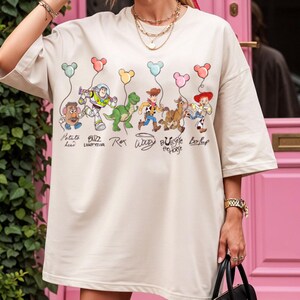May include: Beige oversized t-shirt with a colourful graphic of Toy Story characters holding balloon strings. Characters include Mr. Potato Head, Buzz Lightyear, Rex, Woody, Bullseye the Horse, and Bo Peep. The shirt has short sleeves.
